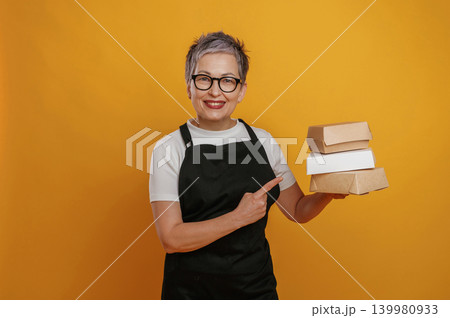 Smiling woman with short gray hair wearing glasses holds stacked takeout boxes against a vibrant yellow background, showcasing food packaging and delivery options Smiling woman with short gray hair wearing glasses holds stacked takeout boxes against a vibrant yellow background, showcasing food packaging and delivery options 139980933