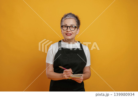 Mature woman with short gray hair wearing glasses and black apron is smiling while holding a notebook and pen against a vibrant yellow background, showcasing creativity and warmth Mature woman with short gray hair wearing glasses and black apron is smiling while holding a notebook and pen against a vibrant yellow background, showcasing creativity and warmth 139980934