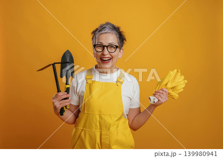 Senior woman gardener in bright yellow overalls smiles while holding gardening tools and gloves against a vibrant yellow background, showcasing gardening enthusiasm 139980941