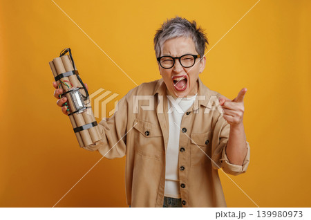 Excited woman with short gray hair holding dynamite sticks, expressing intense emotions against a vibrant yellow background, showcasing a dramatic and bold concept 139980973