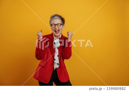 Joyful senior woman in red blazer celebrates success with raised fists against a vibrant yellow background, expressing happiness and excitement in a lively atmosphere Joyful senior woman in red blazer celebrates success with raised fists against a vibrant yellow background, expressing happiness and excitement in a lively atmosphere 139980982