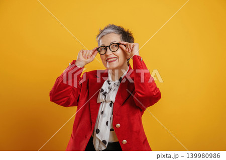 Senior woman with stylish gray hair and glasses is playfully adjusting her eyewear against a vibrant yellow background, showcasing confidence and personality Senior woman with stylish gray hair and glasses is playfully adjusting her eyewear against a vibrant yellow background, showcasing confidence and personality 139980986