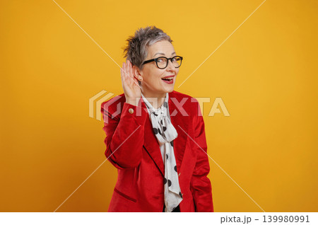 Senior woman with stylish short hair wearing red blazer and glasses, playfully listening with hand to ear against vibrant yellow background, expressing curiosity and engagement Senior woman with stylish short hair wearing red blazer and glasses, playfully listening with hand to ear against vibrant yellow background, expressing curiosity and engagement 139980991