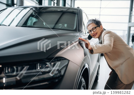 Smiling woman in stylish suit is admiring sleek modern car in bright showroom, showcasing automotive design and elegance with a touch of sophistication Smiling woman in stylish suit is admiring sleek modern car in bright showroom, showcasing automotive design and elegance with a touch of sophistication 139981282
