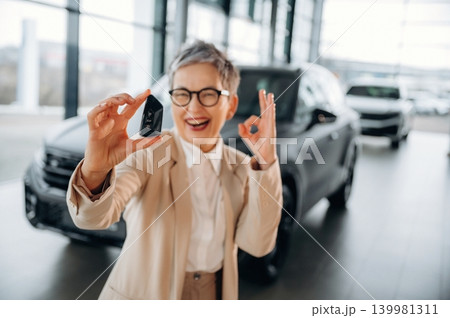 Happy senior woman in stylish outfit is holding car key and showing OK gesture in modern car dealership, celebrating successful vehicle purchase experience Happy senior woman in stylish outfit is holding car key and showing OK gesture in modern car dealership, celebrating successful vehicle purchase experience 139981311