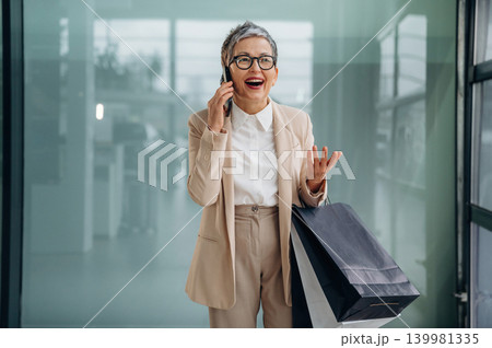 Senior woman in stylish suit is joyfully talking on the phone while holding shopping bags, showcasing a vibrant shopping experience in a modern setting 139981335