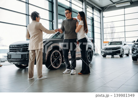 Couple is shaking hands with a car dealer in a modern showroom, showcasing a sleek vehicle and bright, spacious environment with large windows Couple is shaking hands with a car dealer in a modern showroom, showcasing a sleek vehicle and bright, spacious environment with large windows 139981349