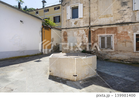 A stone well sits in the middle of a sun-drenched courtyard between weathered buildings. The scene is an outdoor view of a narrow street of Piran, Slovenia 139981400
