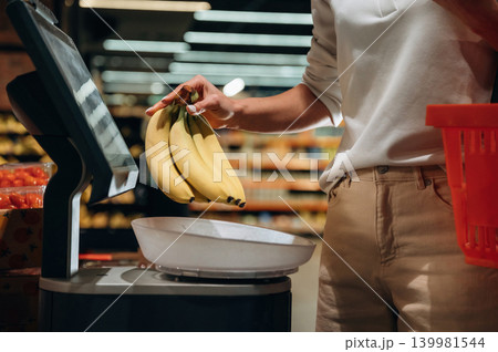 Individual weighing fresh bananas on a digital scale in a grocery store, surrounded by vibrant produce and shopping baskets, showcasing healthy food choices 139981544
