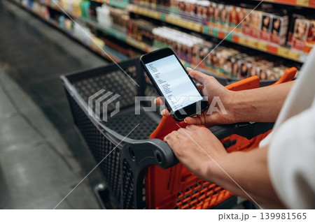 Individual holding smartphone while shopping in a grocery store, with shopping cart and colorful product shelves in the background, showcasing modern shopping experience 139981565