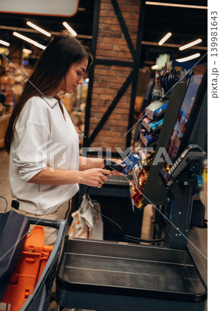 Female cashier scanning items at self-checkout station in a modern grocery store, showcasing the convenience of automated shopping experience Female cashier scanning items at self-checkout station in a modern grocery store, showcasing the convenience of automated shopping experience 139981643