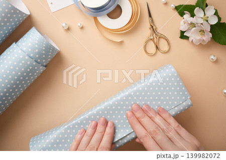 Top view of woman's hands wrapping a gift in blue polka dot paper, surrounded by gold scissors, ribbons, and flowers on a beige background. Top view of woman's hands wrapping a gift in blue polka dot paper, surrounded by gold scissors, ribbons, and flowers on a beige background. 139982072