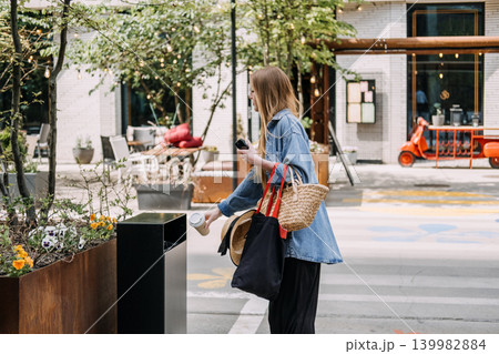 Young woman throws disposable cup into public trash bin on city street. Proper waste management, urban litter control, responsible rubbish disposal. 139982884