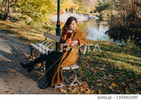 Woman sits on park bench holding travel mug near lake during autumn afternoon. Simple life philosophy outdoor, conscious time management relaxation, appreciation of nature moment. Woman sits on park bench holding travel mug near lake during autumn afternoon. Simple life philosophy outdoor, conscious time management relaxation, appreciation of nature moment. 139982890