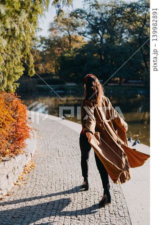Woman walks along paved path by pond in autumn park from behind. Scenic solo vacation exploration, domestic tourism lakeside escape, peaceful weekend travel route. Woman walks along paved path by pond in autumn park from behind. Scenic solo vacation exploration, domestic tourism lakeside escape, peaceful weekend travel route. 139982891