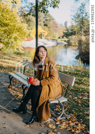 Woman sits on park bench holding travel mug near lake during autumn afternoon. Simple life philosophy outdoor, conscious time management relaxation, appreciation of nature moment. Woman sits on park bench holding travel mug near lake during autumn afternoon. Simple life philosophy outdoor, conscious time management relaxation, appreciation of nature moment. 139982892