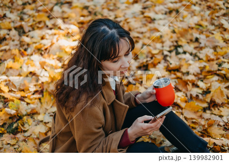 Person sits on ground using smartphone while holding thermal cup in park filled with fallen leaves. Checking banking app, using mobile navigation tool, smartphone online shopping. 139982901