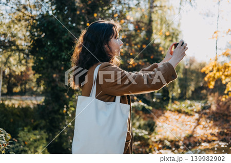 Woman stands on pedestrian bridge holding smartphone to record video of autumn landscape. User generated content strategy, woman capturing marketing video, mobile phone content creation. 139982902