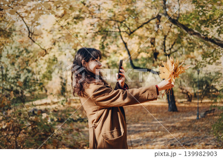 Person takes picture of yellow autumn leaves with smartphone in sunny park. Taking photo with smartphone, mobile photography outdoor, female content creator. 139982903