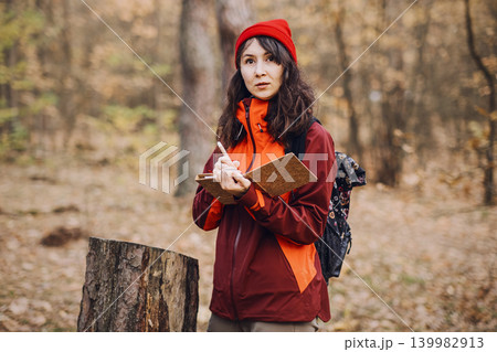 Middle aged woman writes in notebook while standing in autumn forest next to tree stump. Eco-therapy practice, emotional balance outdoors, stress reduction nature. 139982913