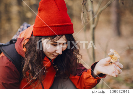 Woman squats in forest holding small wild mushroom for examination. Mushroom identification guide, amateur mycology study, botanical field research, fungal species recognition. Woman squats in forest holding small wild mushroom for examination. Mushroom identification guide, amateur mycology study, botanical field research, fungal species recognition. 139982941