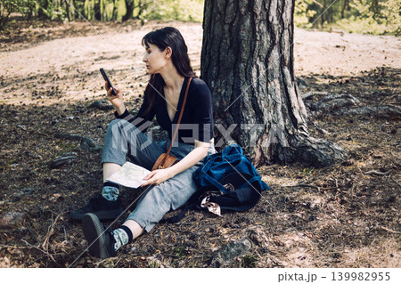 Person sits against tree holding smartphone and paper map in woodland. Backpacking destination guide, nature travel experience, eco-tourism adventure. Person sits against tree holding smartphone and paper map in woodland. Backpacking destination guide, nature travel experience, eco-tourism adventure. 139982955