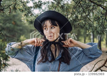 Person wearing hat stands under pine tree branches in forest and adjusts bow on collar. Conscious consumerism lifestyle, ethical fashion brand, eco-friendly apparel. 139982969