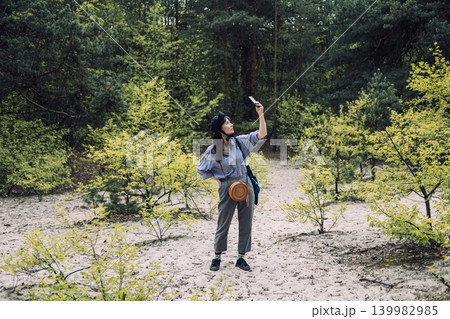 Young woman stands in sandy forest clearing while holding smartphone high to find signal. Remote area connectivity, finding signal in nature, mobile navigation hiking, Digital nomad lifestyle. Young woman stands in sandy forest clearing while holding smartphone high to find signal. Remote area connectivity, finding signal in nature, mobile navigation hiking, Digital nomad lifestyle. 139982985