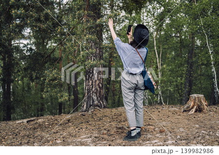 Person wearing hat stands in forest and uses smartphone to identify pine needles on tree. AI plant recognition app, digital botany identifier, real-time nature scanning. Person wearing hat stands in forest and uses smartphone to identify pine needles on tree. AI plant recognition app, digital botany identifier, real-time nature scanning. 139982986