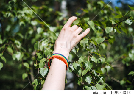 Hand wearing rainbow bracelet reaches toward green birch leaves in forest. Human rights organization, volunteering for LGBTQ cause, community support program. Hand wearing rainbow bracelet reaches toward green birch leaves in forest. Human rights organization, volunteering for LGBTQ cause, community support program. 139983018