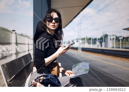 Woman sits on bench at railway platform holding paper map and smartphone. Trip organization, holiday destination search, travel route mapping 139983022