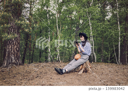 Person wearing hat sits on tree stump in forest and looks at smartphone. AI chatbot for everyday use, virtual assistant in nature, smart AI lifestyle. Person wearing hat sits on tree stump in forest and looks at smartphone. AI chatbot for everyday use, virtual assistant in nature, smart AI lifestyle. 139983042