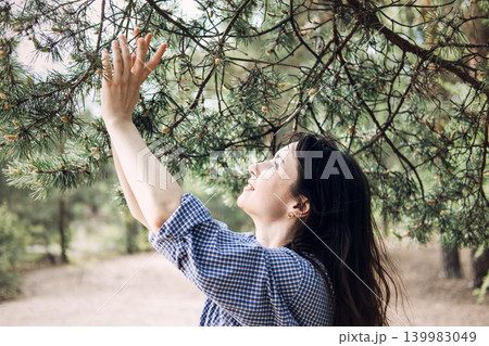 Woman touches pine tree branches while smiling in forest. Nature-based healing, feeling of freedom, psychological benefits of forest Woman touches pine tree branches while smiling in forest. Nature-based healing, feeling of freedom, psychological benefits of forest 139983049