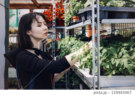 Woman selects leafy green vegetable plants from metal shelf at outdoor garden market. Food self-sufficiency, homegrown produce movement, urban agriculture lifestyle. 139983067