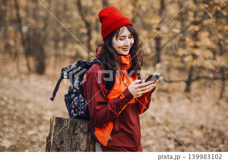 Smiling woman looks at smartphone screen while leaning on tree stump in autumn forest. AI-driven wilderness assistance, mobile tools for trail safety, digital outdoor survival guide. 139983102