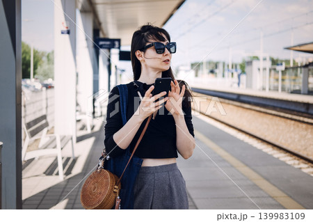 Young woman holds smartphone while standing on sunny railway platform. Modern railway commute, public transport network, sustainable urban travel, Waiting for train. 139983109