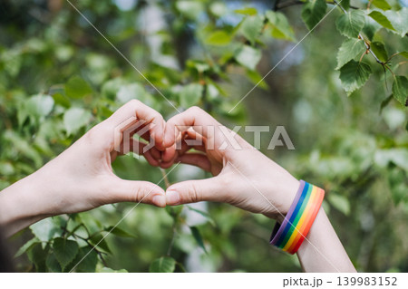 Hands form heart shape against green foliage with rainbow bracelet on wrist. Pride month celebration, LGBTQ pride support, queer community solidarity. 139983152