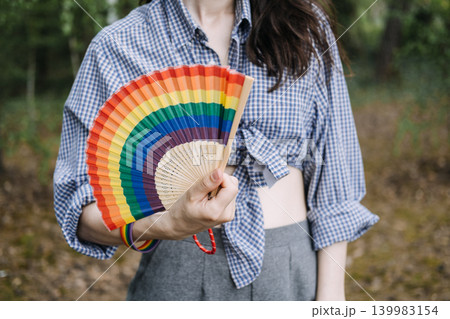 Person holds rainbow folding fan and wears matching wristband while standing outdoors. LGBTQ pride, Queer culture representation, pride parade outfit elements, symbolic rainbow apparel. Person holds rainbow folding fan and wears matching wristband while standing outdoors. LGBTQ pride, Queer culture representation, pride parade outfit elements, symbolic rainbow apparel. 139983154