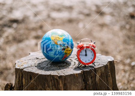 Small globe and alarm clock stand on tree stump against blurred natural background. Saving the earth, climate action now, sustainability and planet protection. Small globe and alarm clock stand on tree stump against blurred natural background. Saving the earth, climate action now, sustainability and planet protection. 139983156