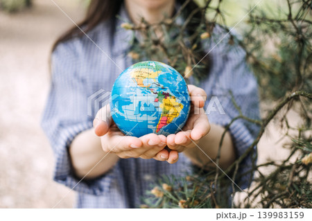 Person holds small globe with both hands outdoors near pine branches. Human touch for nature, protecting global ecosystem, Earth day celebration. 139983159
