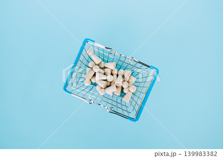 Small wooden peg dolls fill miniature wire shopping basket on bright blue surface. Cost of living crisis, inflation impact on families, basic needs affordability, Consumer purchasing power. Small wooden peg dolls fill miniature wire shopping basket on bright blue surface. Cost of living crisis, inflation impact on families, basic needs affordability, Consumer purchasing power. 139983382