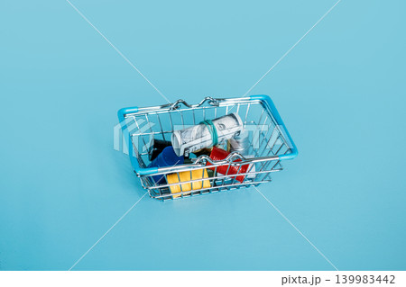 Small shopping basket contains rolled dollar bill resting on miniature oil drums. Economic recession impact, industrial resource consumption, energy market volatility. 139983442
