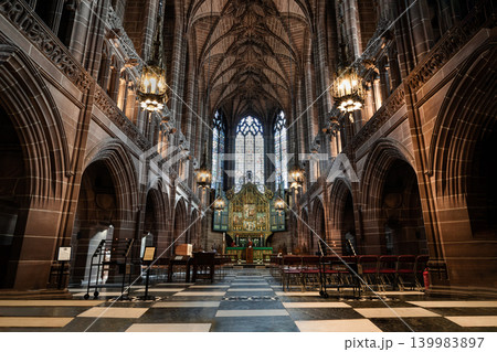 Warm light from hanging lanterns illuminates the towering stone arches and intricate vaulted ceiling of a grand, historic cathedral, casting a serene glow down a checkered aisle toward a magnificent 139983897