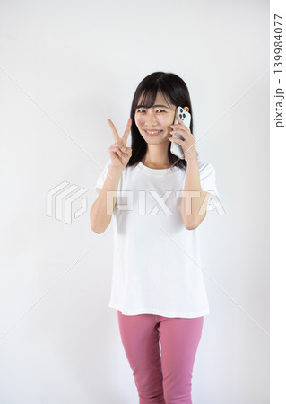 白い背景の前でスマートフォンで通話しながらピースサインをする日本人の若い女性 白い背景の前でスマートフォンで通話しながらピースサインをする日本人の若い女性 139984077