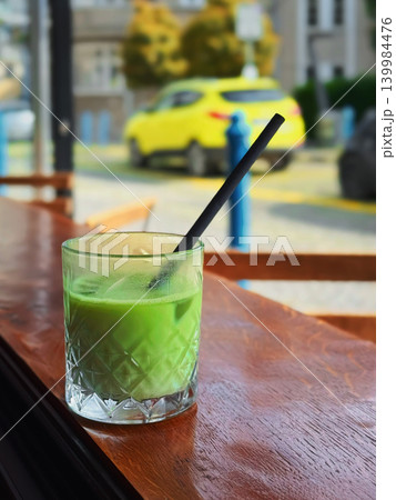 Iced matcha latte in patterned glass with straw stands on wooden cafe table by window with city street blur, copy space. Concept of matcha lifestyle, slow living, cafe break, wellness drink Iced matcha latte in patterned glass with straw stands on wooden cafe table by window with city street blur, copy space. Concept of matcha lifestyle, slow living, cafe break, wellness drink 139984476