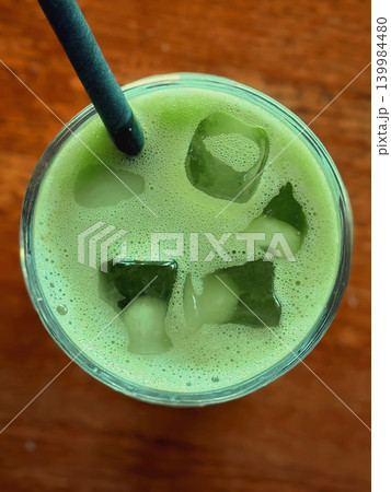 Top view of foamy matcha drink with ice cubes and craft straw in glass on wooden table, green bubbles and chilled surface. Concept of matcha texture, cafe refreshment, wellness beverage trend 139984480
