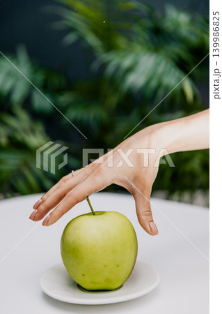 Close-up of a young woman taking a green apple from a plate, with a lush green tree in the background. Healthy lifestyle, fresh fruit, nutrition, wellness, and outdoor concept. Natural ingredients and 139986825