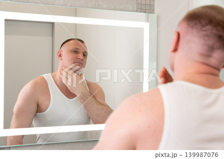 Handsome young man looking at his reflection in bathroom mirror after shaving. Male grooming, skincare and personal care routine concept with clean smooth skin. 139987076