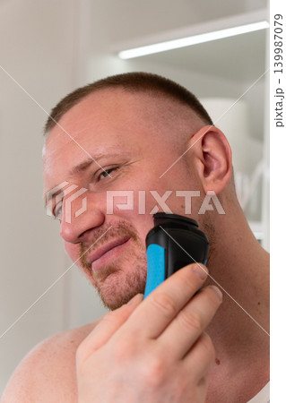 Vertical close up portrait of handsome young man shaving his face with electric shaver in bathroom mirror during morning routine. Male grooming, skincare and personal care concept. 139987079