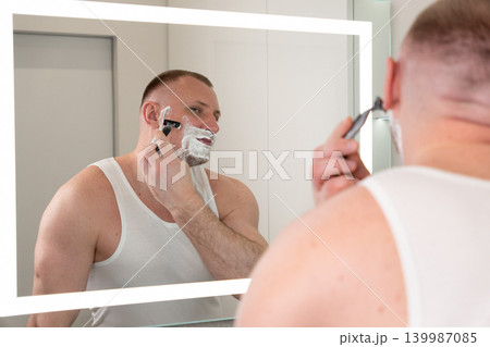 Handsome young man shaving his face with razor in bathroom mirror during morning routine. Male grooming, skincare and personal care concept. Handsome young man shaving his face with razor in bathroom mirror during morning routine. Male grooming, skincare and personal care concept. 139987085